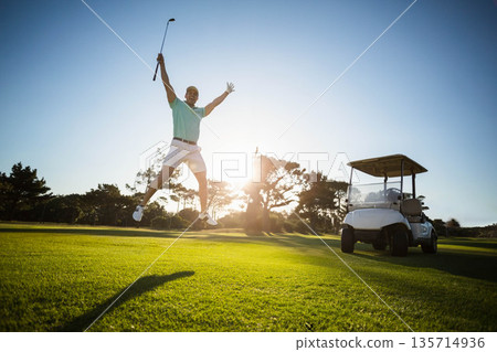 Male golfer jumping raising golf club wearing golf glove on fairway at sunset with golf cart 135714936