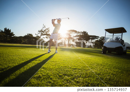 Senior male golfer swinging golf club on fairway during sunset wearing golf attire with golf cart Senior male golfer swinging golf club on fairway during sunset wearing golf attire with golf cart 135714937