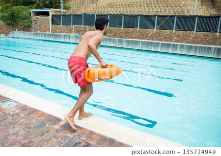 Mid-adult male lifeguard standing poolside holding rescue buoy by lanes in red trunks, copy space Mid-adult male lifeguard standing poolside holding rescue buoy by lanes in red trunks, copy space 135714949