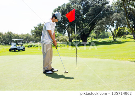 Asian man aligning putter with golf ball on green by red flag with golf cart behind Asian man aligning putter with golf ball on green by red flag with golf cart behind 135714962