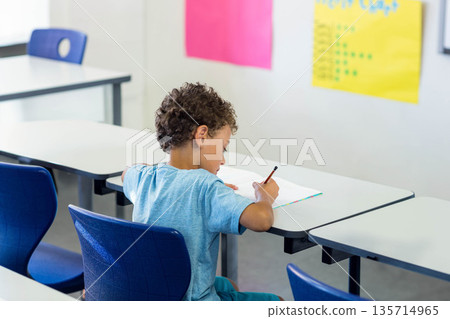 Elementary schoolboy writing in open notebook with pencil in classroom among desks chairs posters 135714965