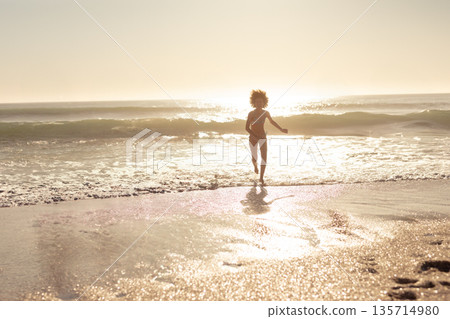 African American woman in bikini running along beach with ocean waves and sun reflection 135714980