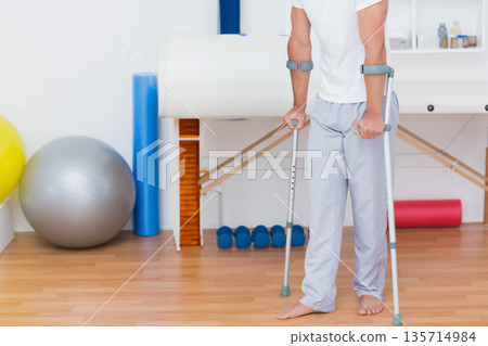 Male patient using forearm crutches standing barefoot in clinic with exercise balls, copy space 135714984