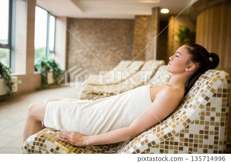 Woman wearing towel reclining on mosaic-tiled lounge chair at spa with large windows, copy space Woman wearing towel reclining on mosaic-tiled lounge chair at spa with large windows, copy space 135714996