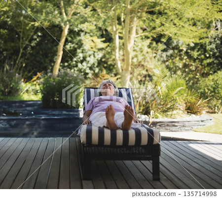 Senior woman relaxing on lounge chair on deck in backyard garden beside reflective water feature 135714998