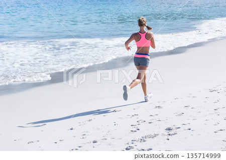 Woman jogging on sandy beach in pink bra, gray shorts, white shoes by ocean, copy space 135714999