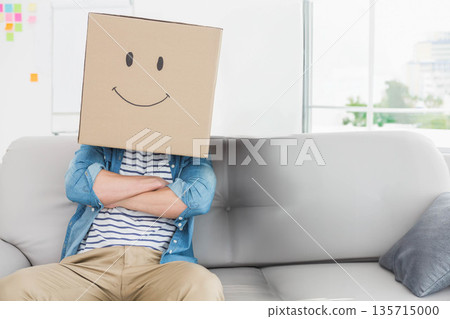 Adult male sitting on gray couch wearing cardboard box head with drawn smile at office lounge Adult male sitting on gray couch wearing cardboard box head with drawn smile at office lounge 135715000