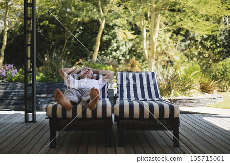 Senior man resting hands behind head on striped lounge chair on deck beside stone planter 135715001