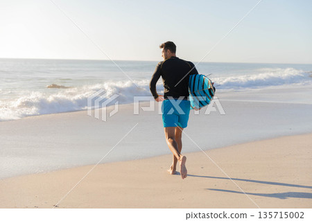 Adult man running along sunlit shoreline at beach carrying striped bodyboard under arm, copy space 135715002