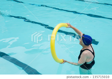 Senior woman gripping yellow ring, wearing goggles and standing waist-deep in lap pool, copy space 135715010