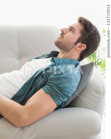 Man reclining on light-colored sofa leaning against gray cushion in living room with potted plant 135715013