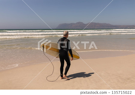 Senior man standing on sandy beach wearing wetsuit holding yellow surfboard with leash facing ocean 135715022