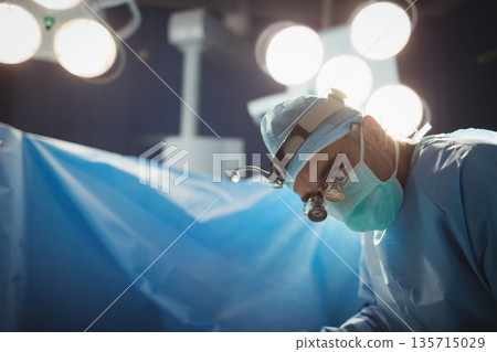 Male gowned surgeon in OR using loupe leaning over draped patient under lights, copy space 135715029