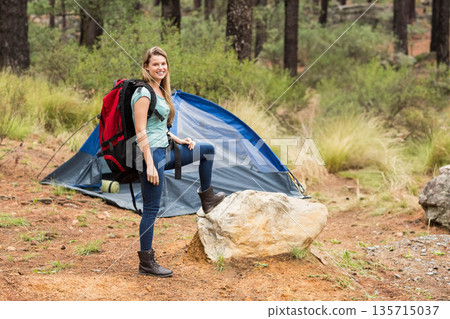 Female hiker carrying red backpack standing beside blue tent near boulder in pine forest clearing Female hiker carrying red backpack standing beside blue tent near boulder in pine forest clearing 135715037