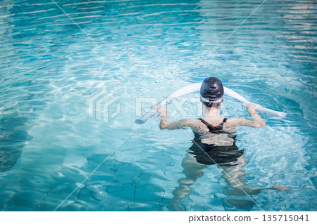 Woman standing chest-deep in outdoor pool wearing swimsuit and cap while holding white foam noodle Woman standing chest-deep in outdoor pool wearing swimsuit and cap while holding white foam noodle 135715041