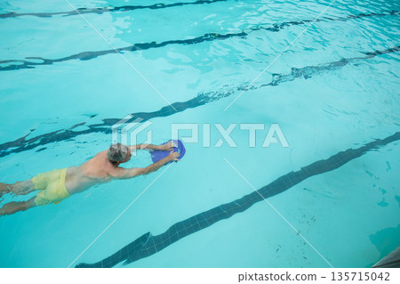 Senior man gliding face-down through lap pool holding blue kickboard with yellow trunks, copy space Senior man gliding face-down through lap pool holding blue kickboard with yellow trunks, copy space 135715042