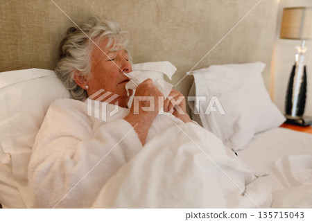 Senior woman reclining on bed wearing white robe blowing nose into tissue beside lamp and pillows Senior woman reclining on bed wearing white robe blowing nose into tissue beside lamp and pillows 135715043