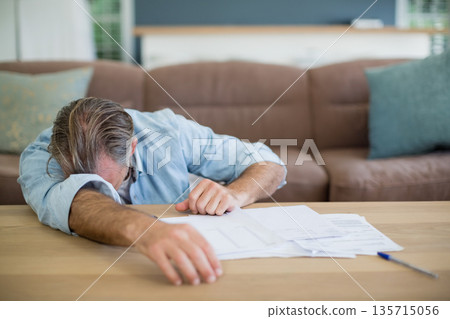 Senior man resting forehead on scattered paperwork and blue pen at home living room coffee table Senior man resting forehead on scattered paperwork and blue pen at home living room coffee table 135715056