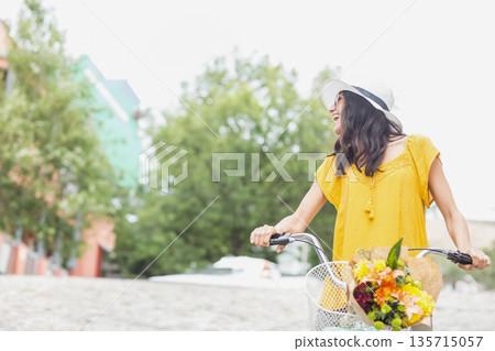 Woman in twenties riding bicycle on cobblestones wearing sunhat carrying flower basket, copy space 135715057