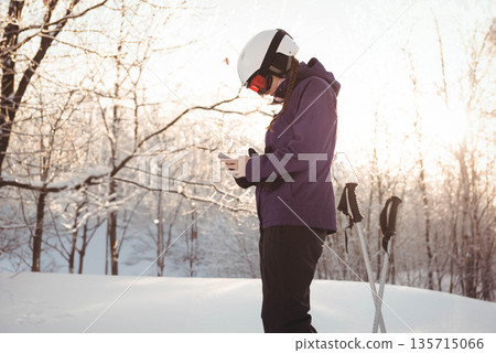 Female teenager standing in snowy clearing checking smartphone with ski poles, copy space 135715066