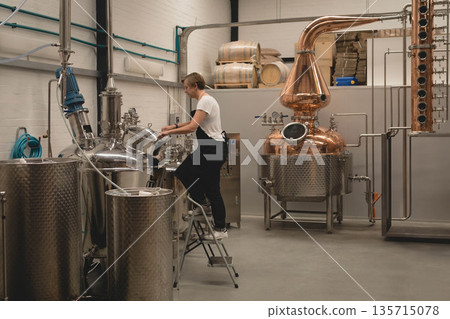 Mature female distillery worker standing on stool adjusting tank valves, copper still at distillery 135715078