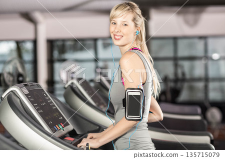 Female exerciser running on treadmill by windows in gym with smartphone and wired earphones 135715079