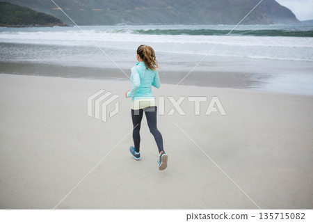Woman jogging on wet sandy beach near ocean waves wearing jacket, leggings and running shoes Woman jogging on wet sandy beach near ocean waves wearing jacket, leggings and running shoes 135715082