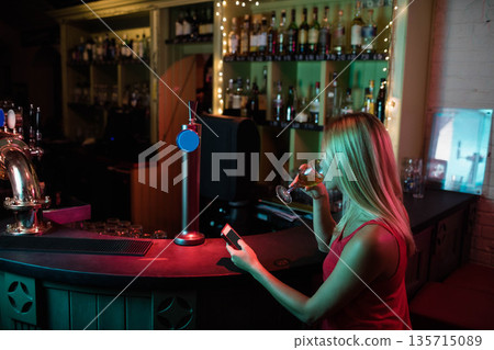 Woman standing at bar under red and green lights holding smartphone and wine glass, copy space 135715089