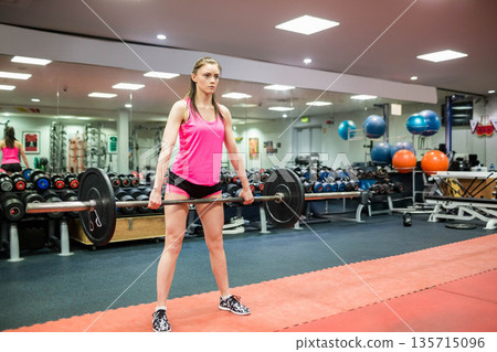 Woman lifting barbell on red mat in fitness center with mirror and exercise balls 135715096