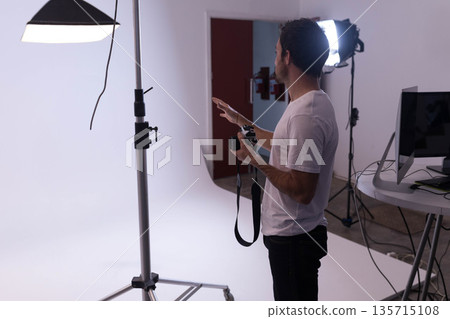 Man holding DSLR camera and gesturing at studio backdrop with softbox light and monitor, copy space 135715108