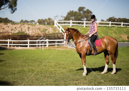 Female equestrian wearing helmet riding chestnut horse with red saddle pad in paddock, copy space Female equestrian wearing helmet riding chestnut horse with red saddle pad in paddock, copy space 135715111