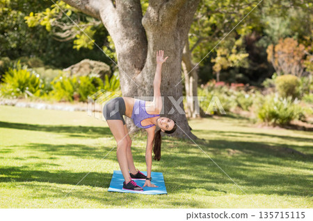 Woman stretching in standing twist on blue mat in park wearing sports bra, shorts, fitness tracker 135715115