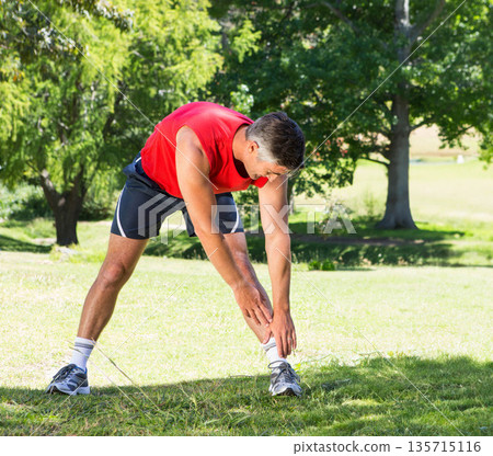 Man standing on lawn in park stretching toward ankles wearing red sleeveless top with running shoes 135715116