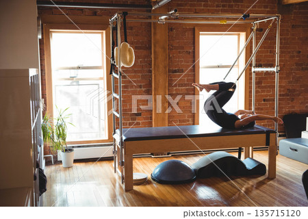 Mid adult woman in black sportswear performing Pilates on reformer in brick walled studio with bosu Mid adult woman in black sportswear performing Pilates on reformer in brick walled studio with bosu 135715120