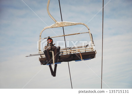 Teenage female skier riding chairlift at ski resort wearing helmet-mounted camera, goggles and skis 135715131