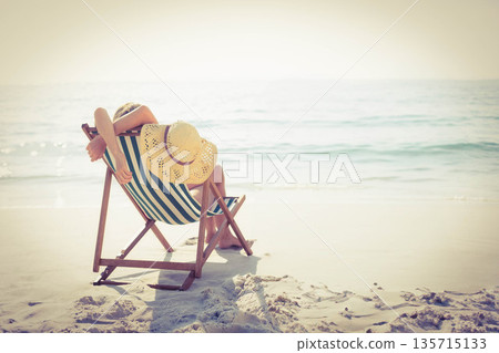 Woman reclining on striped wooden deck chair on white sand seashore, wearing straw sunhat 135715133