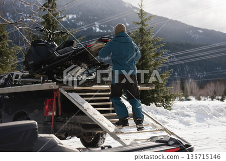 Mid adult man in winter jacket guiding snowmobile up ramp into pickup truck at forest trail Mid adult man in winter jacket guiding snowmobile up ramp into pickup truck at forest trail 135715146