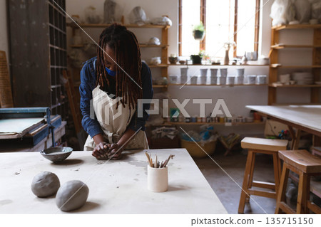 African American woman kneading clay at ceramics workshop table wearing apron with pottery tools African American woman kneading clay at ceramics workshop table wearing apron with pottery tools 135715150