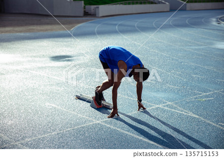 African American adult male athlete aligning in sprint start stance on blue track with metal blocks 135715153