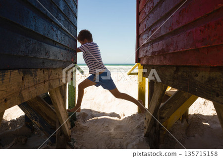 School-age boy stepping between blue and red wooden cabins on sandy beach with ocean waves School-age boy stepping between blue and red wooden cabins on sandy beach with ocean waves 135715158