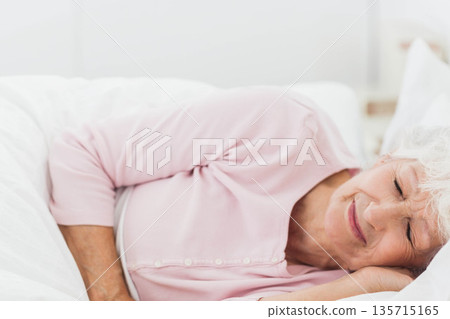 Senior woman lying on side in bed wearing pink pajama top with white pillow and duvet 135715165