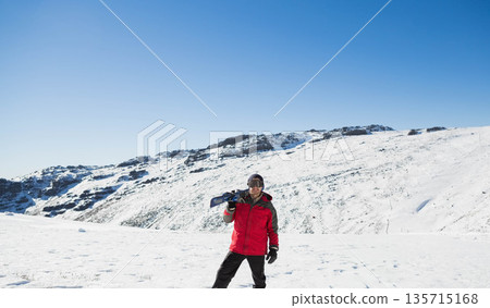 Man standing in snowy valley under blue sky holding snowboard wearing ski goggles, gloves, beanie Man standing in snowy valley under blue sky holding snowboard wearing ski goggles, gloves, beanie 135715168