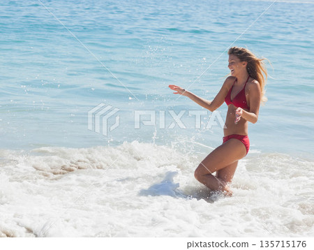Woman standing knee-deep in breaking ocean waves on sandy beach wearing red bikini, copy space Woman standing knee-deep in breaking ocean waves on sandy beach wearing red bikini, copy space 135715176