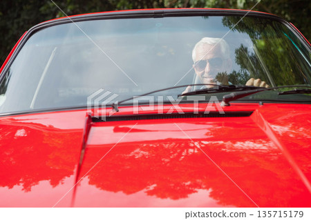 Senior man wearing aviator sunglasses driving red convertible featuring wipers on leafy driveway Senior man wearing aviator sunglasses driving red convertible featuring wipers on leafy driveway 135715179