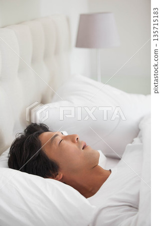 Asian man lying on bed in bedroom with upholstered headboard, white duvet and gray lamp Asian man lying on bed in bedroom with upholstered headboard, white duvet and gray lamp 135715183