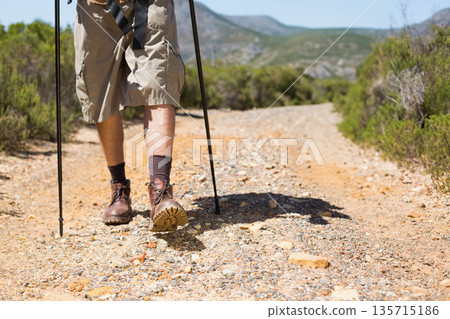 Man hiking along rocky valley path using adjustable trekking poles and wearing cargo shorts Man hiking along rocky valley path using adjustable trekking poles and wearing cargo shorts 135715186
