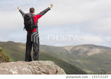 Male hiker raising arms while overlooking mountain panorama on rocky summit with black backpack Male hiker raising arms while overlooking mountain panorama on rocky summit with black backpack 135715187