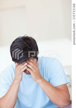 Man sitting in living room wearing light blue t-shirt under soft light covering face with hands 135715188