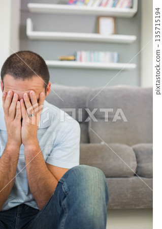 Man leaning on grey couch covering face with hands in living room under shelves, copy space Man leaning on grey couch covering face with hands in living room under shelves, copy space 135715194