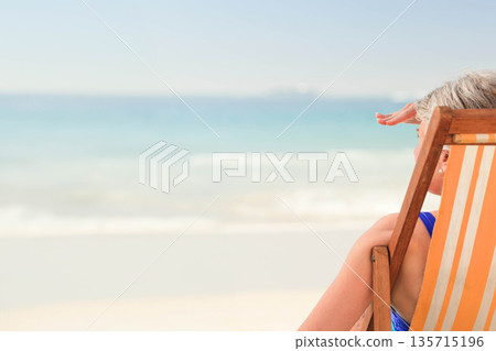 Senior woman sitting in orange-striped beach chair at sandy shore shading eyes from sun, copy space 135715196